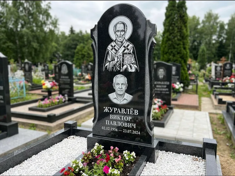 Памятник с гравировкой иконы: стела с образом в полный рост на обороте — гравировка образа Матроны Московской, низкие кусты спиреи в цвету, работа «Лебедева» в Ижевске
